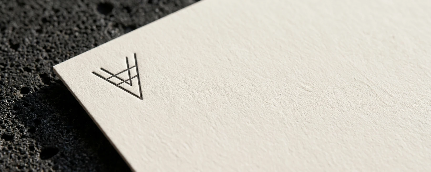 Extreme close-up of blind-debossed V monogram on 350gsm cotton card stock under raking daylight